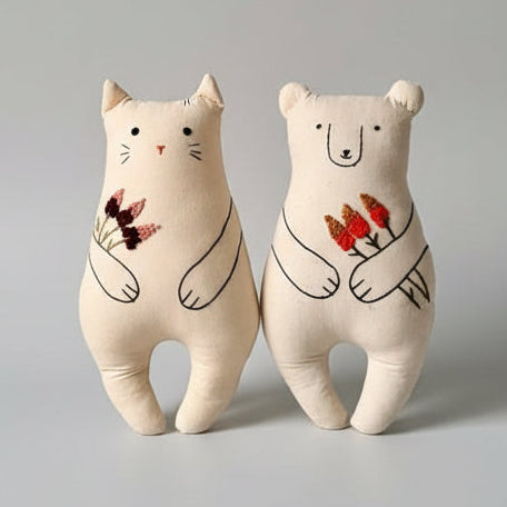 Two plush toys, a cat and a bear, holding flowers against a plain background