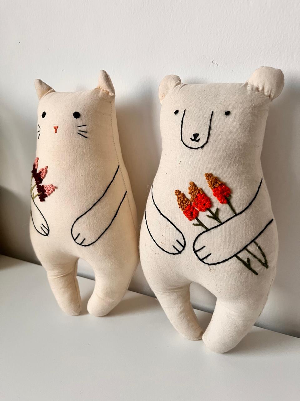 Two plush toys, a cat and a bear, holding flowers against a plain background